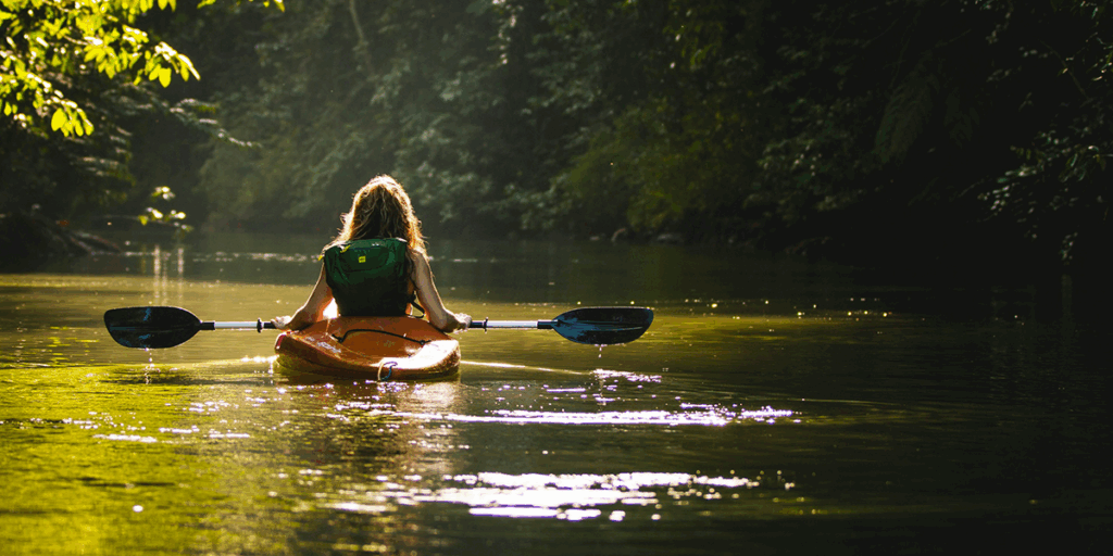 girl riding alone in boa, in the river that is covered with the trees and green leaves from boath side and the sun reflects on the surface of water, a full depiction of searching oneself.