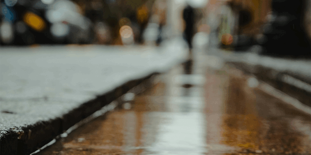 A wet road and the pouring rain, a man can be seen in the rain, walking of the road, with the blurred umbrella in his hands. why get soaked when you already have an umbrella.