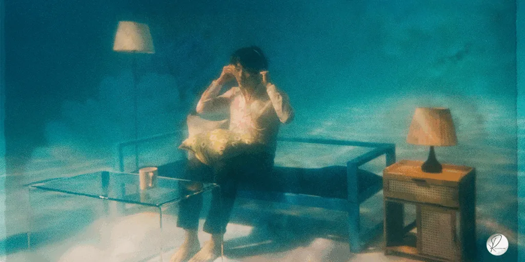 A person sitting at a desk under water with a bed, table, mug, and headphones, symbolizing reflection, self-observation, and gradual personal growth. Represents how ordinary people quietly work on their habits, focus, and productivity while navigating everyday life.